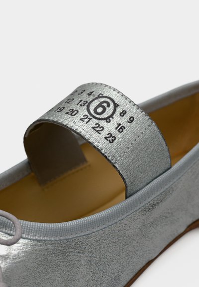 Ασημένιο επίπεδο παπούτσι με λουράκι που φέρει τυπωμένους αριθμούς και έναν μεγάλο αριθμό 6 μέσα σε κύκλο στο πάνω μέρος του λουριού.