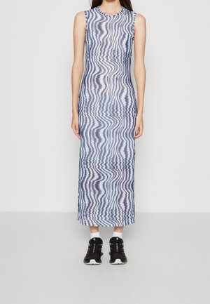 Frau trägt ein ärmelloses, knöchellanges Kleid mit blau-weißen wellenförmigen Streifen, kombiniert mit schwarzen Turnschuhen und weißen Socken, steht vor einer weißen Wand.