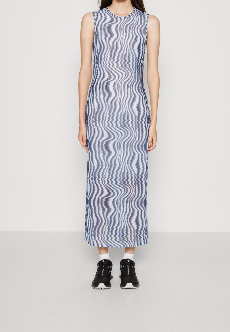 Femme portant une robe sans manches, longue jusqu'aux chevilles, avec des rayures ondulées bleues et blanches, assortie à des baskets noires et des chaussettes blanches, debout contre un mur blanc.