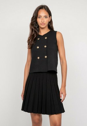 Femme aux longs cheveux foncés portant un haut noir sans manches avec des boutons dorés et une jupe plissée noire assortie, debout devant un fond uni.