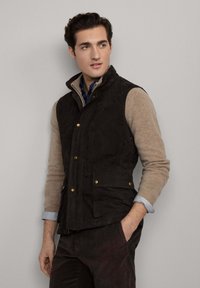Gilet trapuntato marrone scuro con hardware dorato, indossato sopra un maglione beige. Presenta un colletto alto e due tasche frontali.