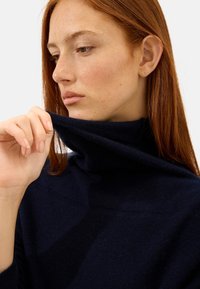 Navyblauer Rollkragenpullover aus weichem Stoff, mit hohem Kragen. Nahaufnahme einer Hand, die den Kragen hält, mit subtilen Texturdetails.