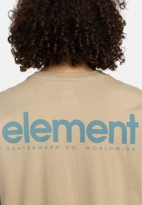 Beige t-shirt med et stort blåt "element" logo på bagsiden, med "SKATEBOARD CO. WORLDWIDE" i mindre tekst under. Lavet af bomuldsstof.