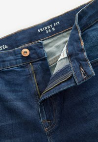 Jean skinny bleu avec un bouton en cuivre et une fermeture éclair. Présente des coutures contrastantes et une étiquette avec la taille 34 R sur la taille.