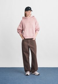 Világos rózsaszín kapucnis pulcsi elülső zsebbel, laza fazonban. Barna bő nadrággal és szürke sportcipővel párosítva. Fekete sapka teszi teljessé a szettet.