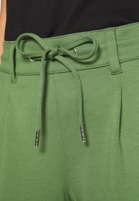 Zelené kalhoty s gumovým pasem a kovovými špičkami, vyrobené z elastického materiálu s hladkou texturou a přizpůsobeným střihem.