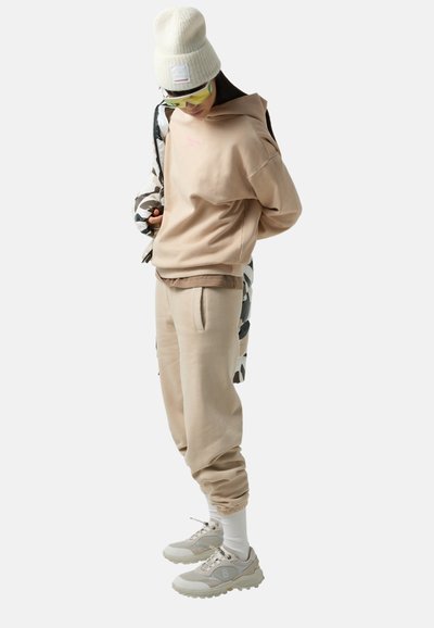 Beżowa, oversize'owa bluza, beżowe dresy i jasne sneakersy. Wełniana czapka i okulary przeciwsłoneczne w zielonym odcieniu. Akcenty w postaci kurtki w kamuflażowy wzór.