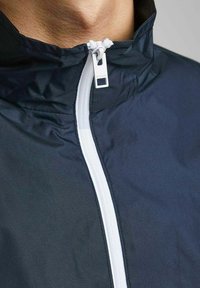 Chaqueta cortavientos navy con cuello alzado, cremallera blanca y tejido suave y texturizado. Vista detallada del cuello y el área de la cremallera.