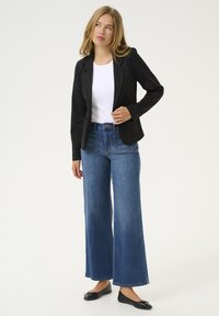 Schwarze Blazer über einem weißen Oberteil, kombiniert mit weit geschnittenen, hoch taillierten blauen Jeans. Das Model trägt schwarze Ballerinas. Sauberes, minimalistisches Design.