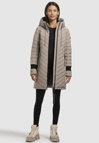 Abrigo largo acolchado beige con capucha, que presenta detalles en negro, un cierre de cremallera y dos bolsillos laterales. Combinado con leggings negros y botas gruesas.