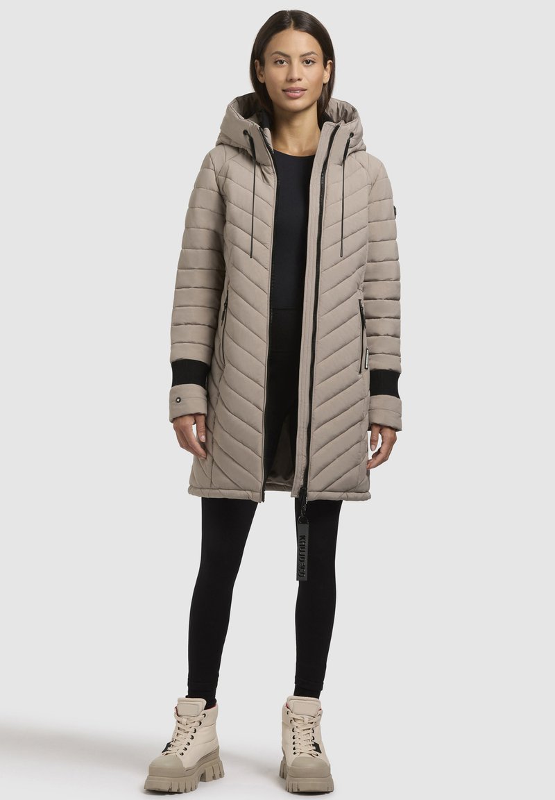 Abrigo largo acolchado beige con capucha, que presenta detalles en negro, un cierre de cremallera y dos bolsillos laterales. Combinado con leggings negros y botas gruesas.