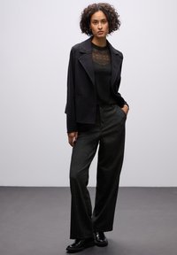 Blazer in lana nera sopra un top con inserti in pizzo nero, abbinato a pantaloni larghi grigio carbone e scarpe lucide nere. Design semplice ed elegante.