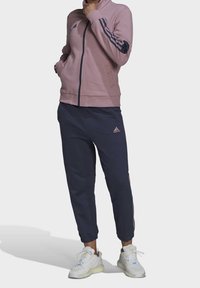 Adidas trainingspak met een roze jas met rits en marineblauwe accenten, gecombineerd met bijpassende marineblauwe joggingsbroek. Gemaakt van zachte stof met elastische manchetten.