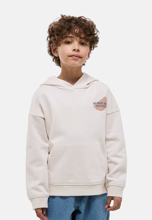 Enfant aux cheveux bouclés portant un sweat à capuche beige clair avec l'inscription "No Bad Days Never" et un jean bleu, debout devant un fond uni.