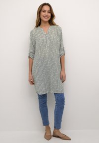 Tunique à motif en vert clair et bleu avec un col en V, manches trois-quarts, associée à un jean bleu ajusté et des ballerines beiges.