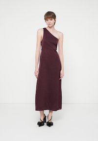 Femeie îmbrăcată într-o rochie midi fără mâneci, cu un umăr, de culoare violet închis, dintr-un material texturat, asortată cu pantofi cu toc înalt negri, cu fundițe.