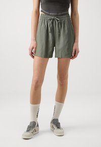 Osoba ubrana w oliwkowe szorty z możliwością regulacji w pasie, szary krótki top, białe skarpetki do kostek z czarnym nadrukiem oraz szare buty Adidas.