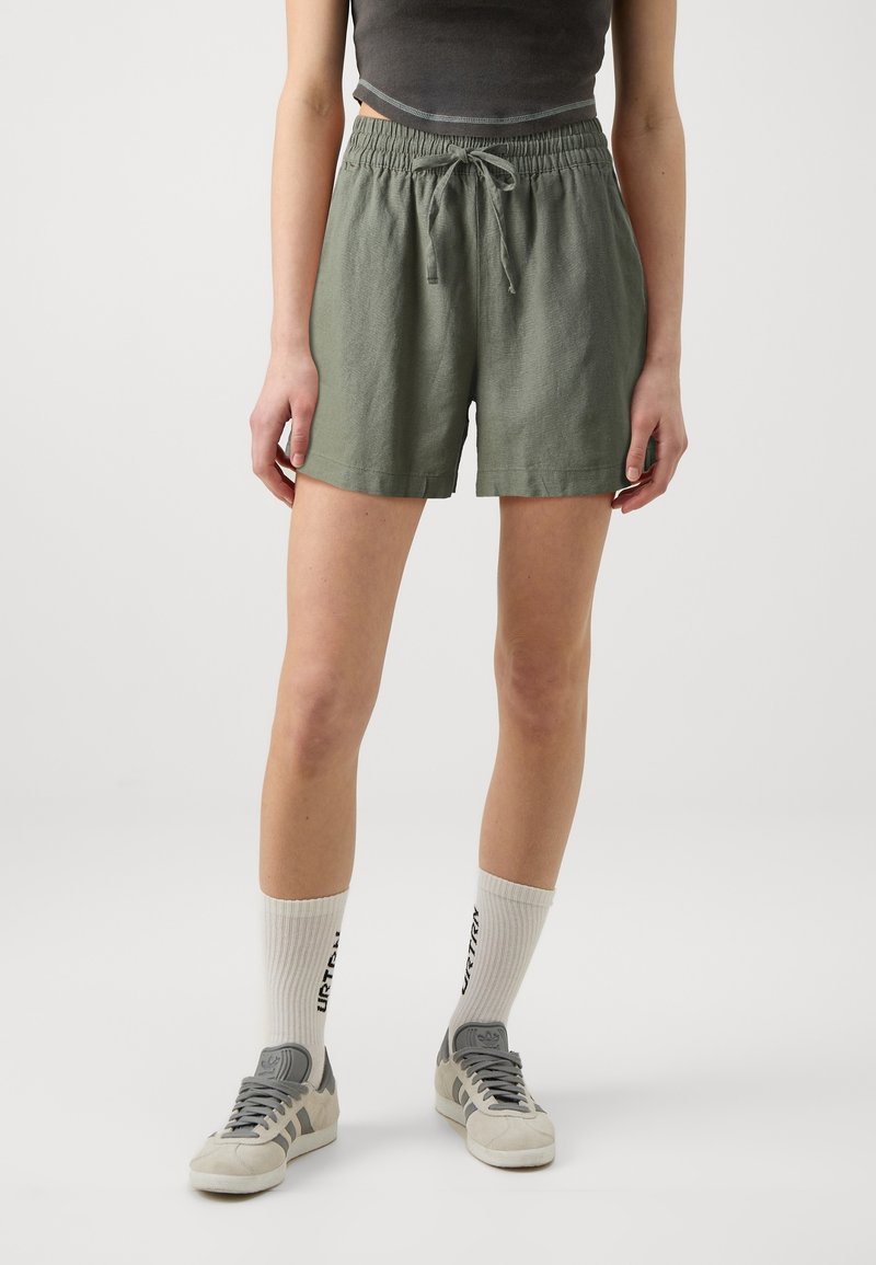 Osoba ubrana w oliwkowe szorty z możliwością regulacji w pasie, szary krótki top, białe skarpetki do kostek z czarnym nadrukiem oraz szare buty Adidas.