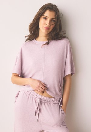 Noor naine laineliste pruunide juustega, seljas lõdva istuv heleroosa lühikeste varrukatega pluus ja sobivad nööriga püksid, seisab ühevärvilise tausta ees.