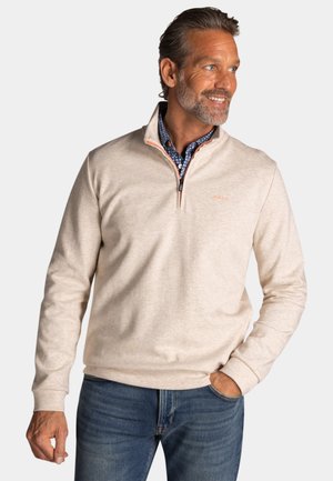 Mann mit kurzen grauen Haaren und Bart trägt beigen Pullover mit kurzem Reißverschluss über blau gemustertem Hemd und blauer Jeans, lächelt und schaut nach rechts.