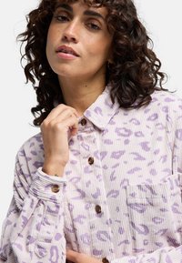 Koszula z kordurowej tkaniny w jasnoróżowym kolorze z purpurowym nadrukiem w panterkę, posiadająca klasyczny kołnierzyk, zapięcie na guziki oraz kieszeń na piersi. Teksturowana tkanina.