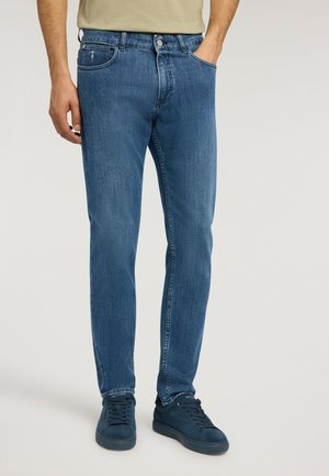 Uomo che indossa jeans blu slim fit e sneaker blu scuro, in piedi con una mano in tasca contro uno sfondo semplice.
