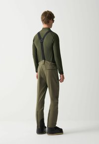 Mann in olivgrünen Skihosen mit schwarzen Hosenträgern, olivgrünem Langarmoberteil und schwarzen Skischuhen, der mit dem Rücken zu einer weißen Hintergrund steht.