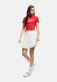 T-shirt rossa cropped con stampa "Malibu Bay California", abbinata a una gonna bianca plissettata. Il modello indossa sneakers bianche e calze a metà polpaccio.
