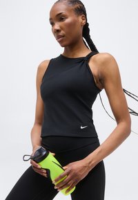 Γυναίκα με πλεξούδες, που φοράει μαύρο αθλητικό τοπ χωρίς μανίκια, κρατώντας ένα φθορίζον πράσινο παγούρι σε ένα απλό φόντο.