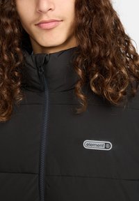 Person med langt krøllet hår iført en sort zip-jakke med et "element" logo på brystet foran en ensfarvet baggrund.