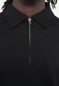 Μαύρο πόλο μπλουζάκι με φερμουάρ στη γραμμή του λαιμού, με απαλή υφή και κλασικό γιακά, που αναδεικνύει έναν μινιμαλιστικό σχεδιασμό.