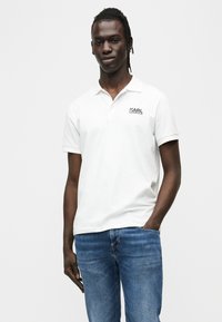 Λευκό βαμβακερό πόλο μπλουζάκι με κοντά μανίκια· διαθέτει μαύρο λογότυπο στο αριστερό στήθος. Φοριέται με μπλε τζιν τζιν. Απλός σχεδιασμός.