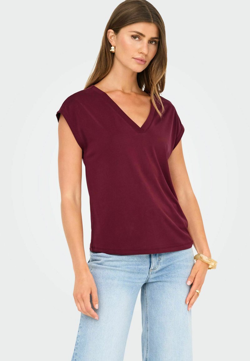 Top de manga corta en burdeos con escote en pico y corte holgado; textura suave; combinado con pantalones vaqueros de mezclilla azul claro, con un fondo neutro.