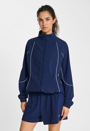 Junge Frau mit lockigem Haar trägt eine marineblaue Jacke mit Reißverschluss und weißen Paspeln sowie passende marineblaue Shorts.