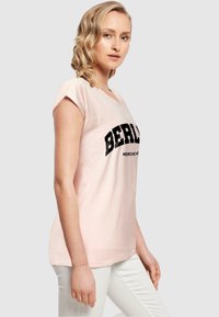 Lyserød V-hals T-shirt med rullede ærmer, der har sort tekst "BERLIN" og "MERCH. CO." nedenunder; lavet af blødt bommateriale.