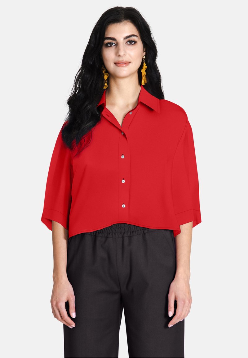 Camicia rossa a maniche corte con bottoni, corta e abbinata a pantaloni neri con vita elastica. Tessuto liscio, colletto semplice e dettagli dei bottoni.