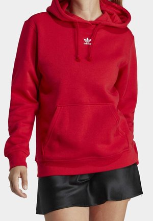 Kobieta w czerwonej bluzie Adidas z kapturem, przednią kieszenią i sznurkami, w połączeniu z czarną mini spódniczką na jednolitym tle.