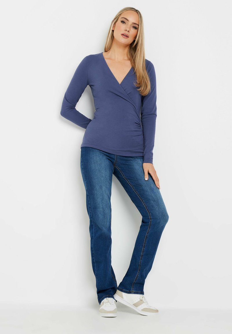 Top a maniche lunghe blu navy con scollatura a V e dettaglio arricciato sul lato. Abbinato a jeans in denim blu e sneakers bianche con accenti beige.