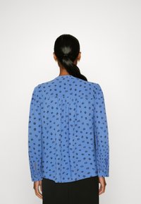 Blaue Bluse mit floralem Muster, langen Ärmeln, gefälteltem Rückendetail und knöpfbaren Manschetten, aus leichtem Material. Schwarze Hose sichtbar.