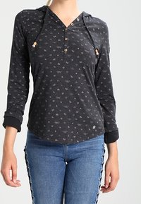 Kvinna i en svart långärmad tröja med huva och små ljusa mönster, samt blå jeans med svarta snörningsdetaljer på sidorna.