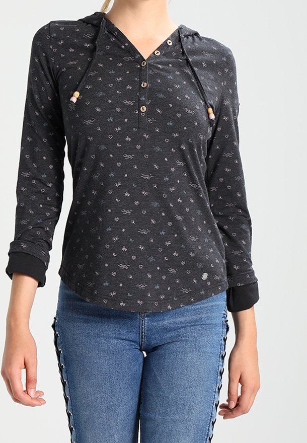 Kvinna i en svart långärmad tröja med huva och små ljusa mönster, samt blå jeans med svarta snörningsdetaljer på sidorna.