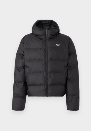 Mężczyzna w czarnej puchowej kurtce z wysokim kołnierzem, z logo Adidas na piersi oraz czarnych dresowych spodniach z białymi pasami po bokach, stojący na jednolitym tle.