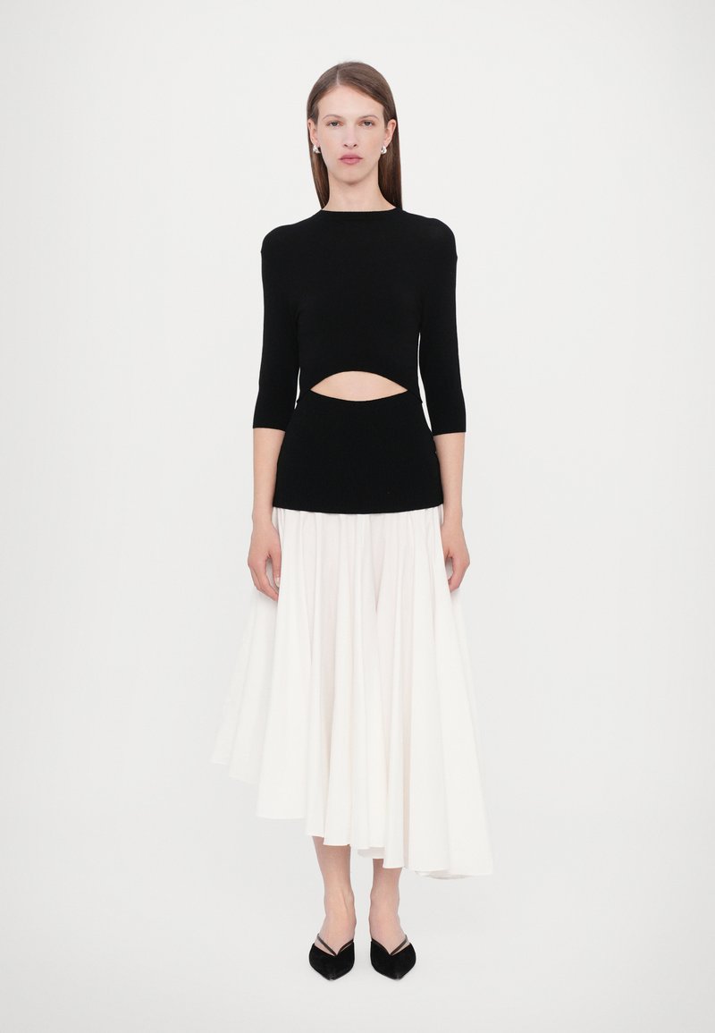 Schwarzes Strickoberteil mit dreiviertel Ärmeln, das ein Cut-out-Detail an der Taille aufweist, kombiniert mit einem fließenden weißen plissierten Rock. Schwarze spitze Ballerinas.