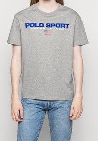 Šedé bavlněné tričko s kulatým výstřihem, krátkými rukávy a výrazným modrým nápisem „POLO SPORT“ doplněným malým motivem americké vlajky.