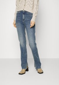 Jeans de mezclilla azul con campana y aberturas en los dobladillos, combinados con un cárdigan de punto beige claro, que presenta un patrón de agujeros en zigzag.