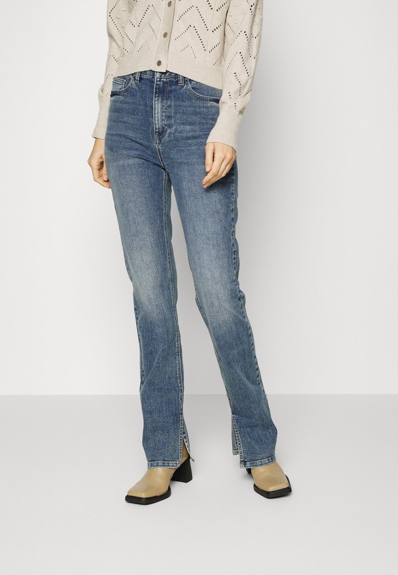 Jeans de mezclilla azul con campana y aberturas en los dobladillos, combinados con un cárdigan de punto beige claro, que presenta un patrón de agujeros en zigzag.