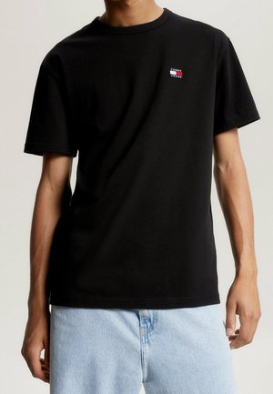 Czarna koszulka z krótkim rękawem wykonana z bawełny, z małym kolorowym logo na lewej piersi. Noszona z jasnoniebieskimi dżinsami.