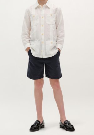 Garçon portant une chemise blanche à manches longues avec quatre poches, un short bleu marine et des mocassins noirs brillants, debout les mains dans les poches.