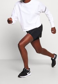 Sudadera blanca con puños acanalados, combinada con pantalones cortos negros. El modelo lleva zapatillas de correr negras con acentos blancos. Pierna en movimiento.
