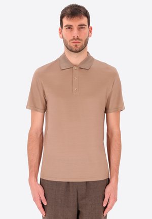 Uomo con capelli corti e scuri e barba, che indossa una polo beige a maniche corte e pantaloni marroni, in piedi davanti a uno sfondo semplice.